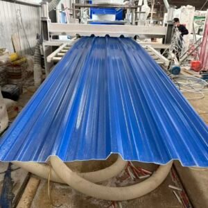 Blue corrugated metal roofing sheet being processed on a conveyor in a busy fabrication shop with a worker in the background wearing a mask.