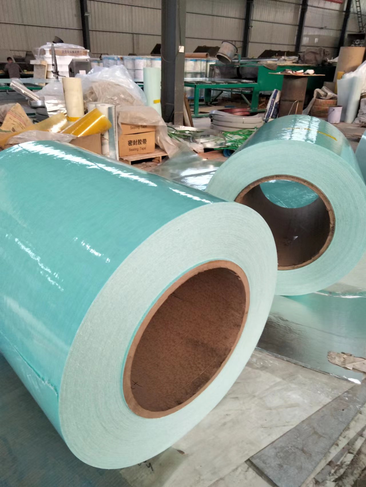 微信图片_20260216231341_137_14 - Large teal insulation rolls in a factory setting, stacked on the floor with cardboard cores visible in a warehouse.
