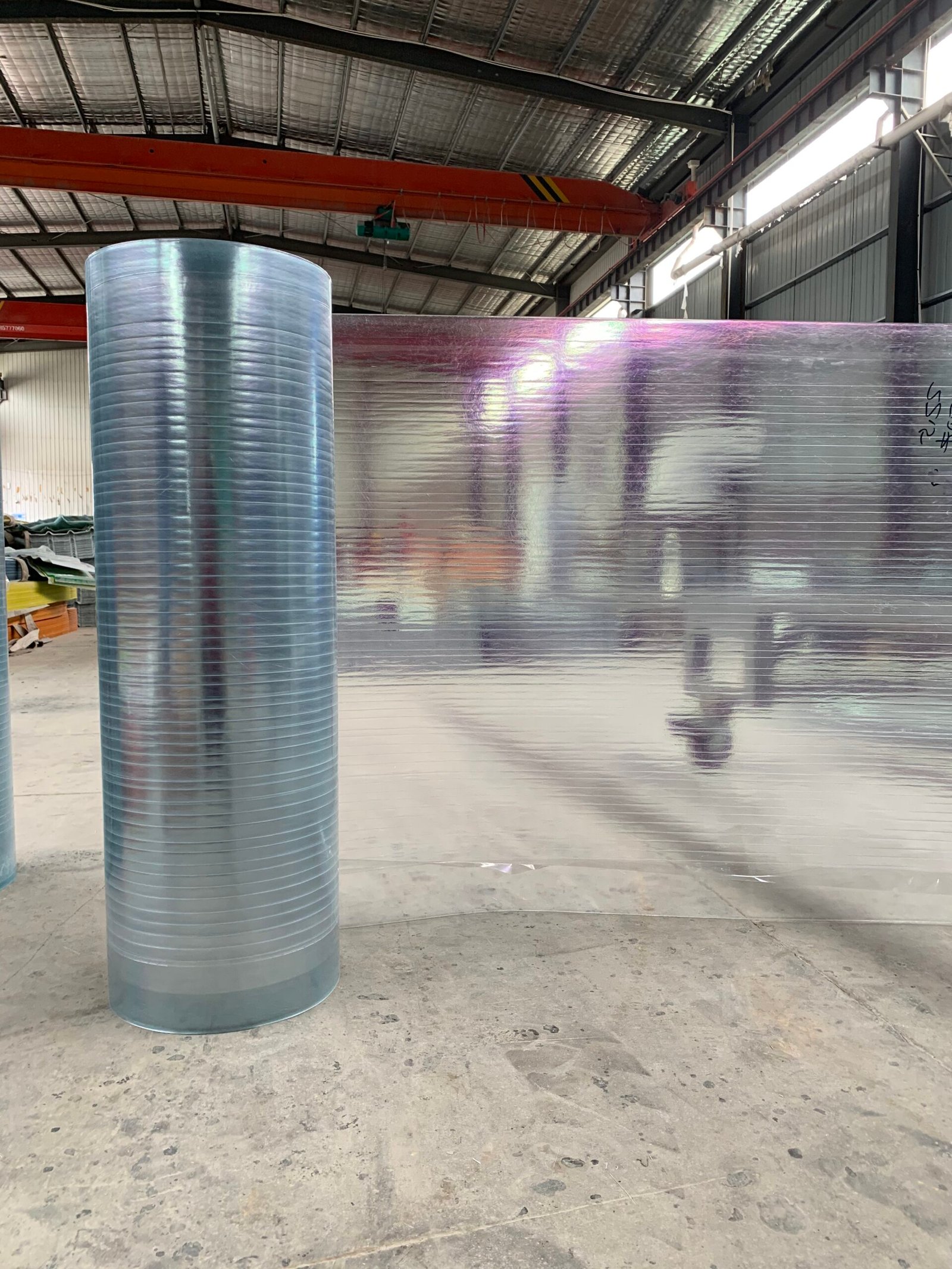 微信图片_20260216231402_141_14 - Industrial warehouse interior with a large blue corrugated metal roll in the foreground and a reflective foil-wrapped wall to the right, under an orange overhead crane.