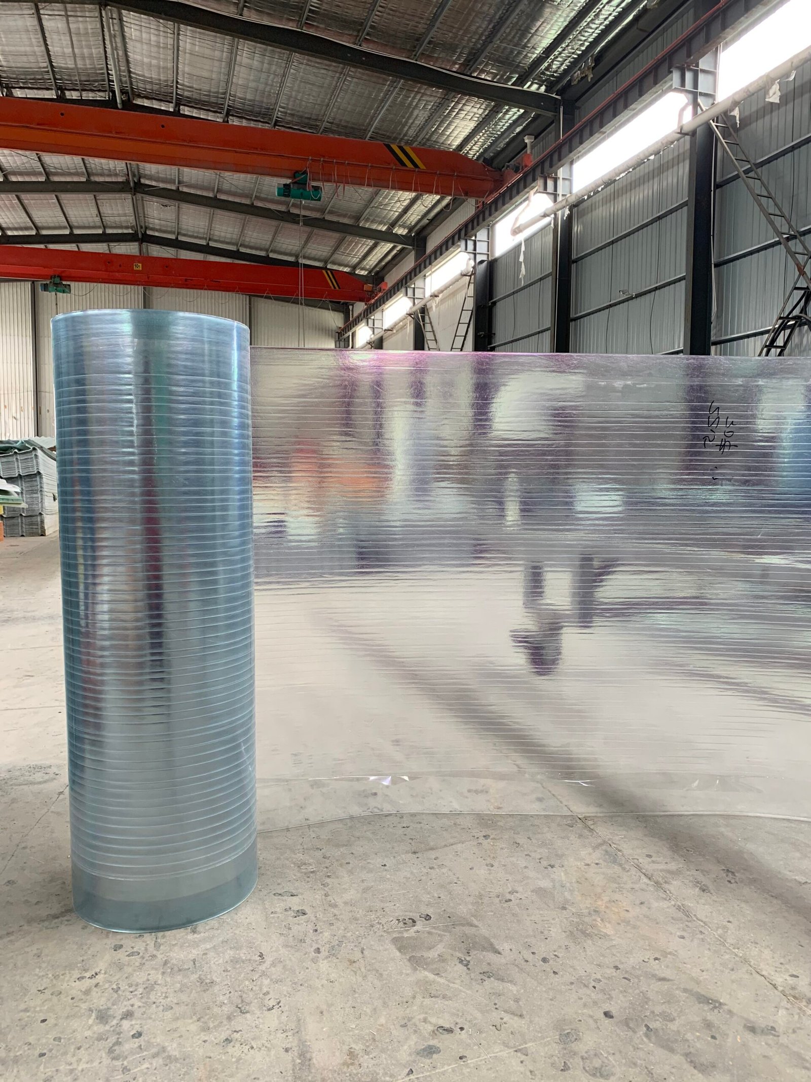 微信图片_20260216231408_142_14 - Inside a factory: a tall blue ribbed roll stands beside a large reflective foil-wrapped sheet, with orange crane beams overhead.