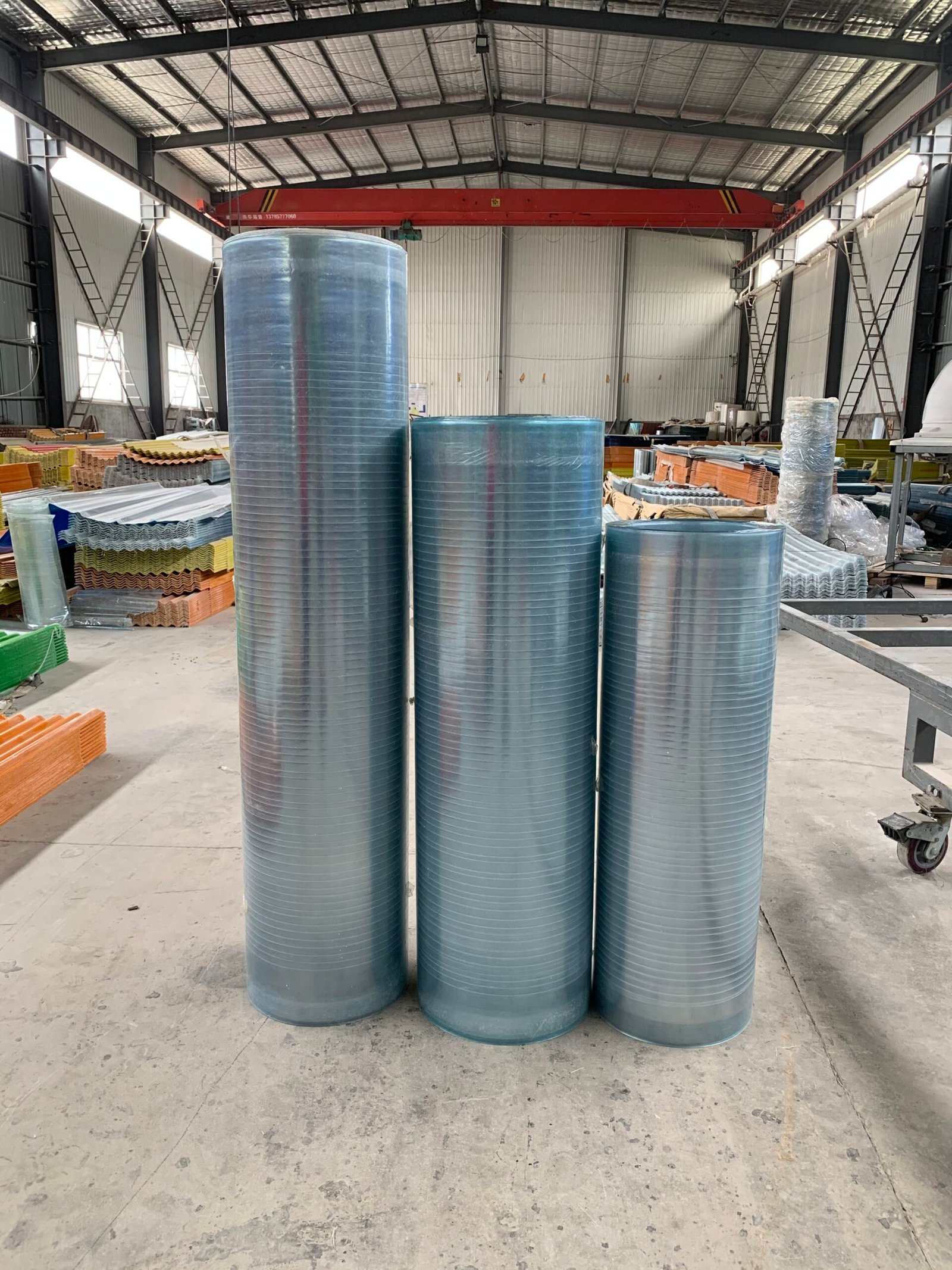 微信图片_20260216231413_143_14 - Three tall blue corrugated metal cylinders standing upright in a warehouse with a red overhead crane and stacked building materials in the background.