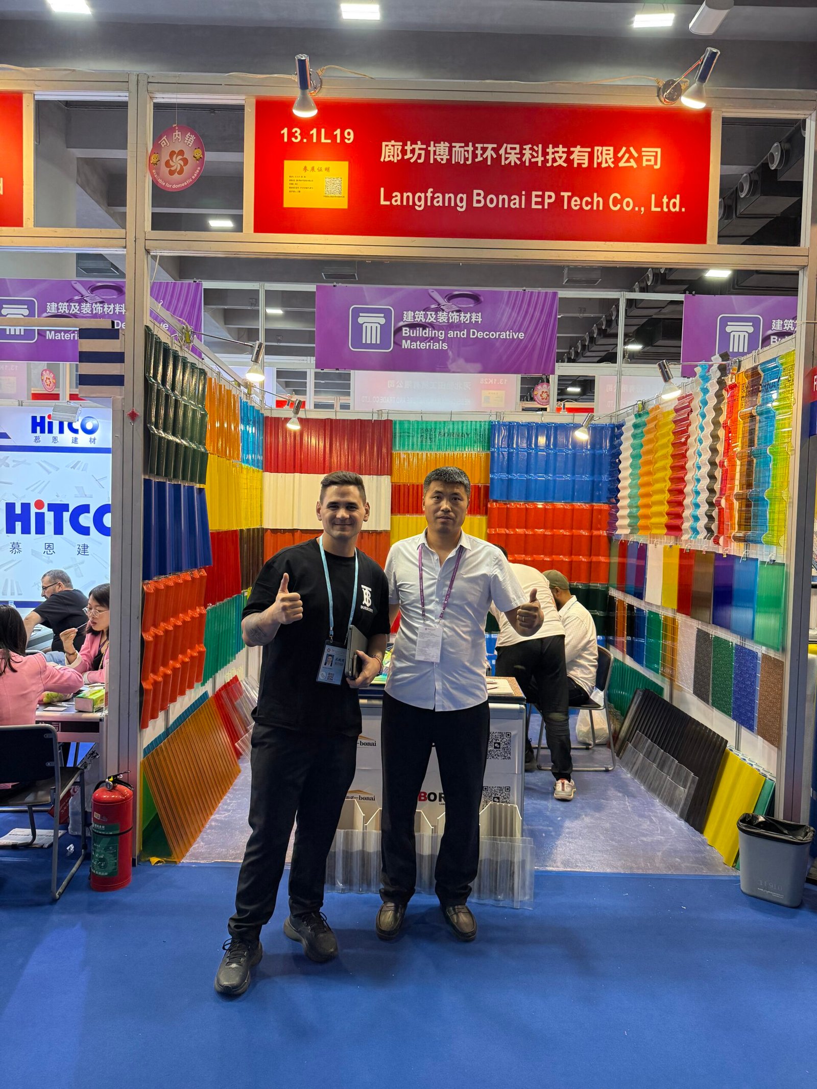 微信图片_20260330144105_440_14 - Trade show booth with colorful plastic panels on display; two men pose and thumbs up in front of the booth banner that reads Langfang Bonai EP Tech Co., Ltd.