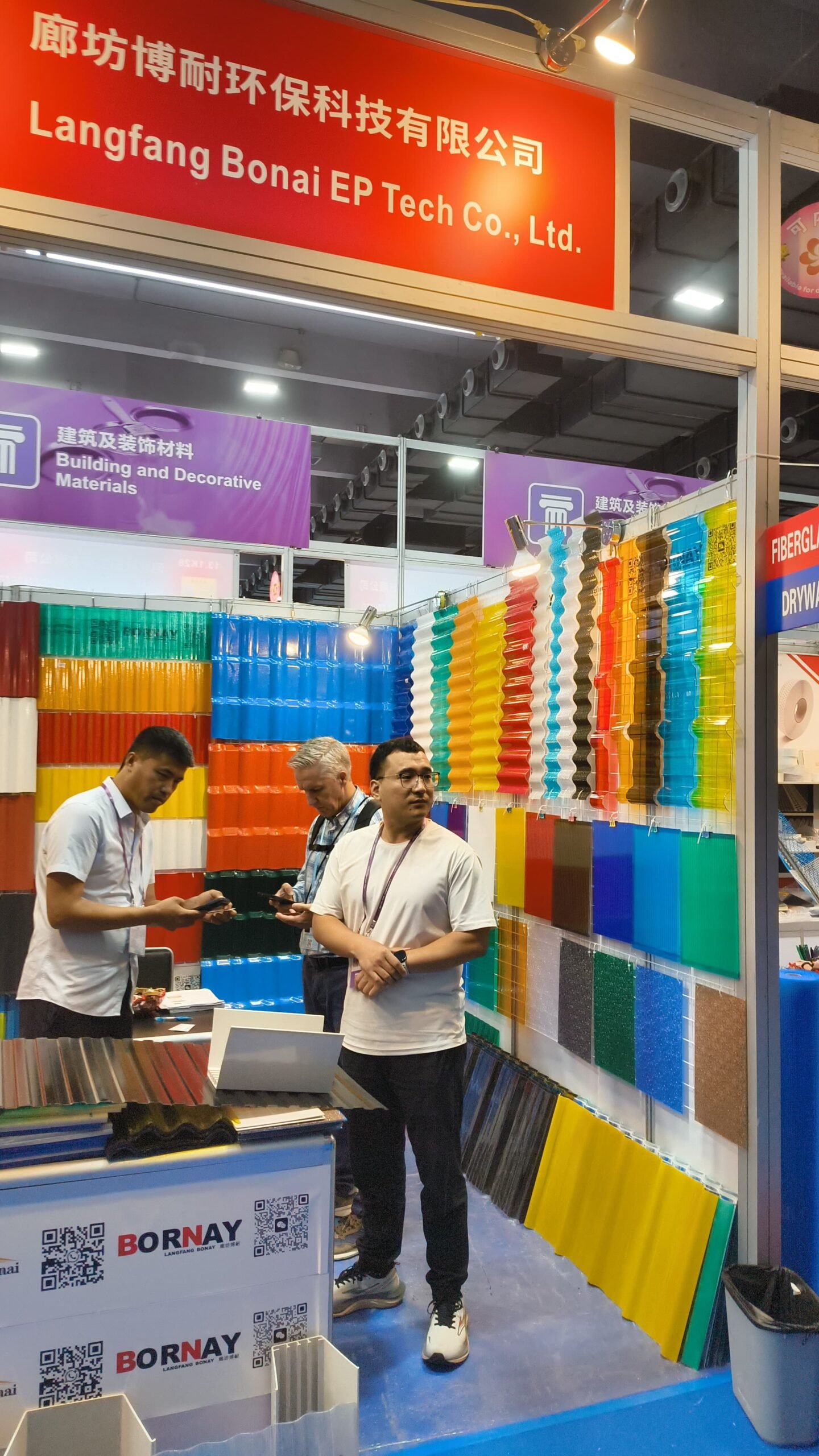 微信图片_20260330144848_448_14 - Trade show booth with a wall of colorful sample panels and three men standing, a red sign above reads Langfang Bonai EP Tech Co., Ltd.