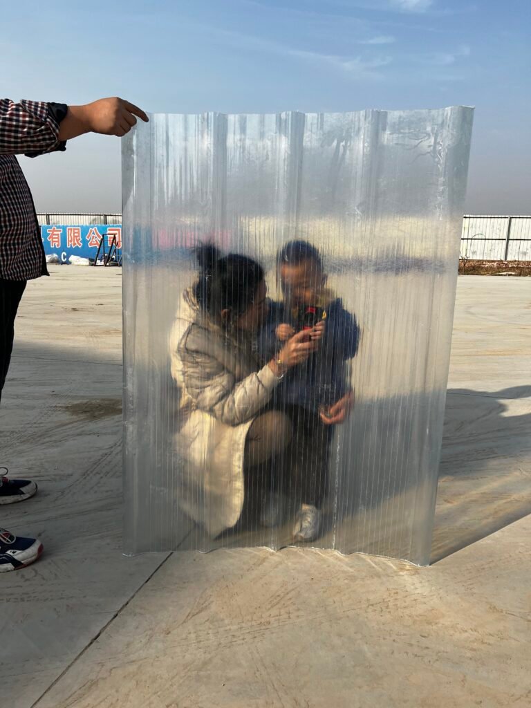 Two people crouch behind a tall clear plastic panel; a hand from the left points toward it in an outdoor setting.