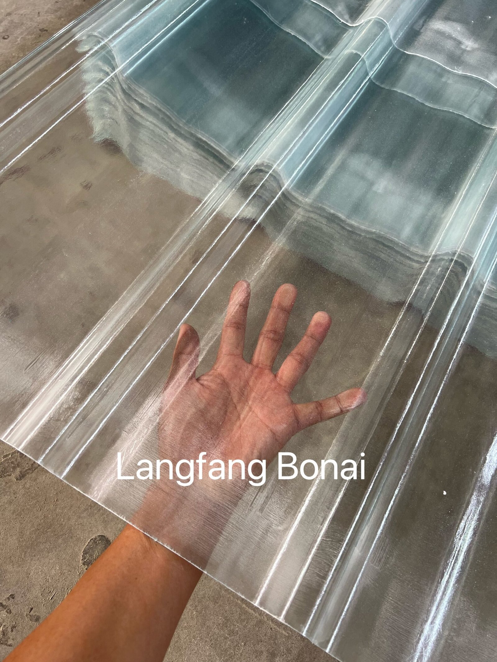微信图片_20260416215257_755_4361 - Person's hand rests on a large clear corrugated plastic sheet laid on a dusty concrete floor, showing seams and reflections.