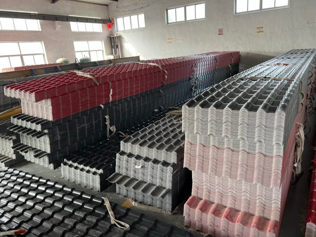 微信图片_20260416215334_782_4361 - Indoor industrial storage of stacked corrugated metal roofing sheets in black, red, and white, strapped and awaiting transport near large windows.