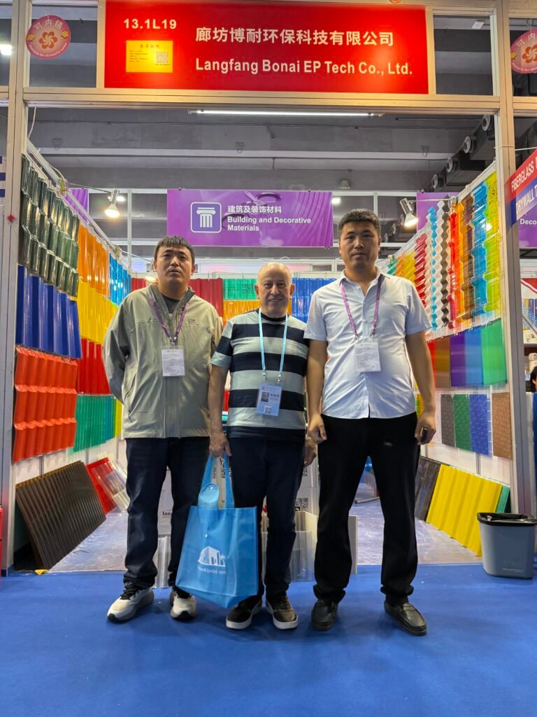 微信图片_20260330144917_456_14 - Three men at a trade show booth surrounded by colorful sample panels; purple sign reads 'Building and Decorative Materials'.