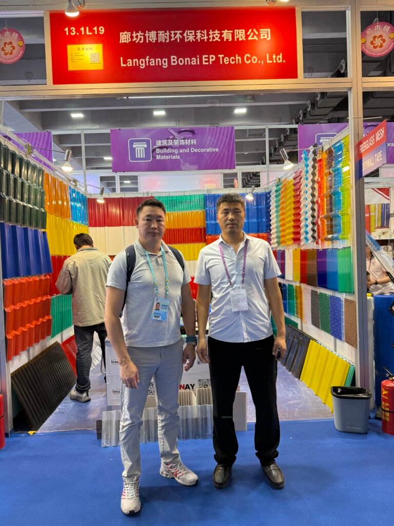 微信图片_20260330145635_457_14 - Two men stand at a trade-show booth displaying rows of colorful plastic sheets and panels under a red sign with Chinese text and company name in the background.